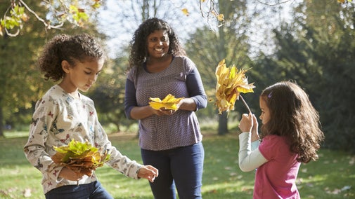 Two children and one woman play with leaves in the early autumn sunshine